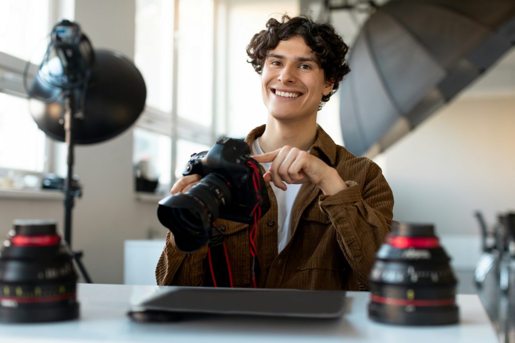 Cheerful male photographer sitting at workplace in modern photostudio, holding professional camera