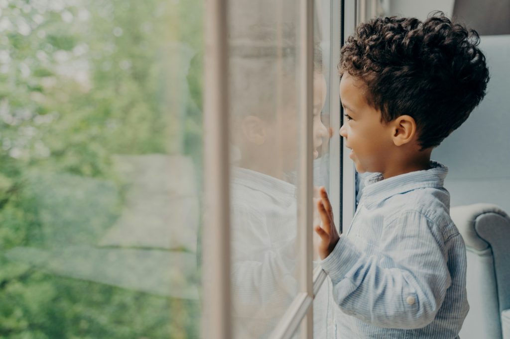 Curious child peers out a window dreamily, enveloped by a soft natural light.