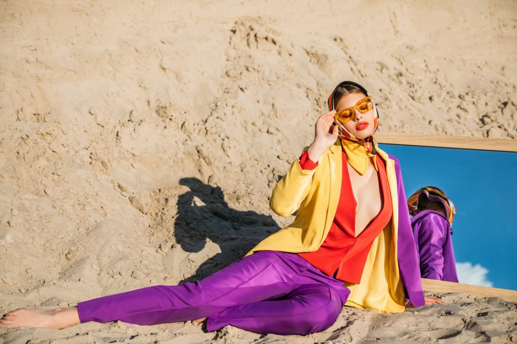 elegant model in fashionable clothes posing near mirror on sand dune