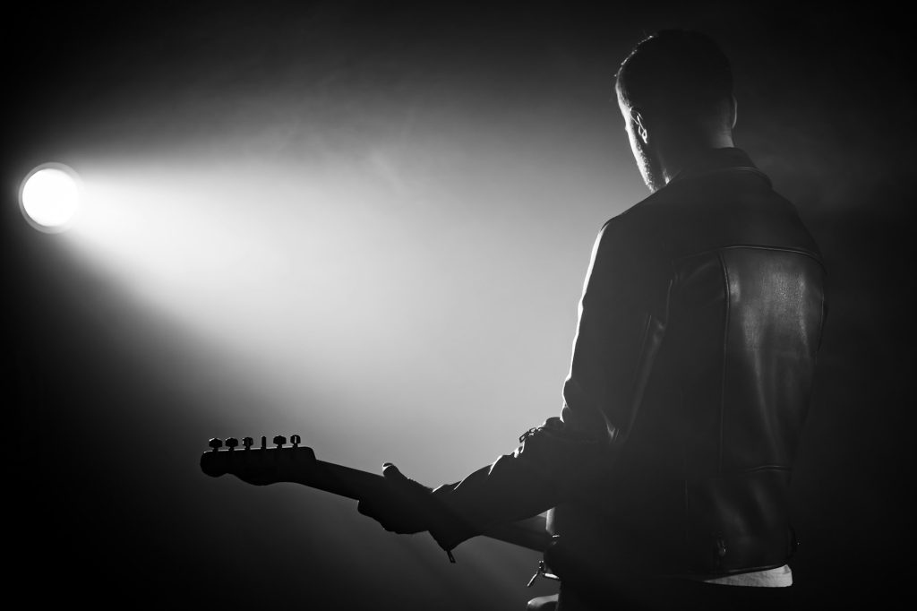 Rock guitarist man in leather jacket standing his back in smoky studio or stage